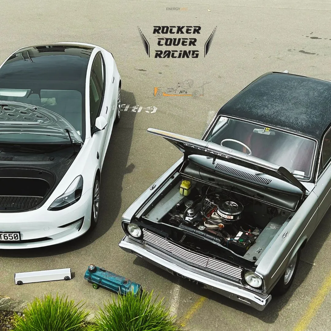 Two cars parked nose in, seen from above. Beside a white modern saloon sits a green classic with its bonnet raised over the engine bay, and on the tarmac in front of them a bare rocker cover and a small blue toy racer.