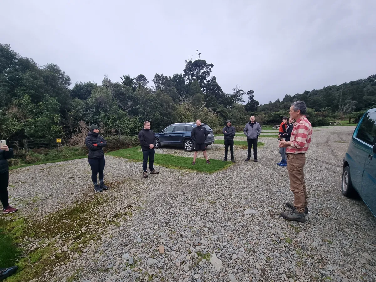 A group standing about on a gravel car park at the edge of native bush under a flat grey sky, their cars parked behind them.