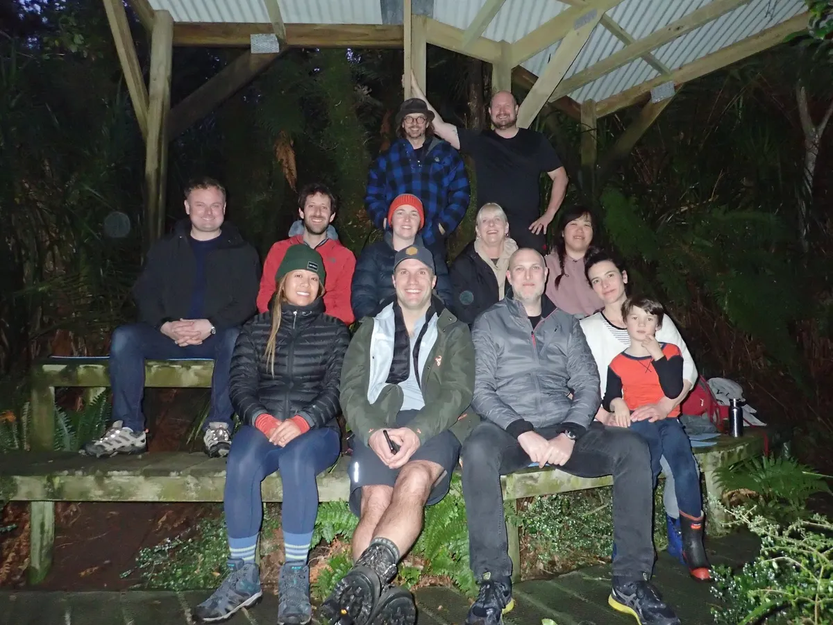 The group gathered under a timber shelter in the bush at night, some seated along a bench and others standing behind, lit by a flash against the dark trees.