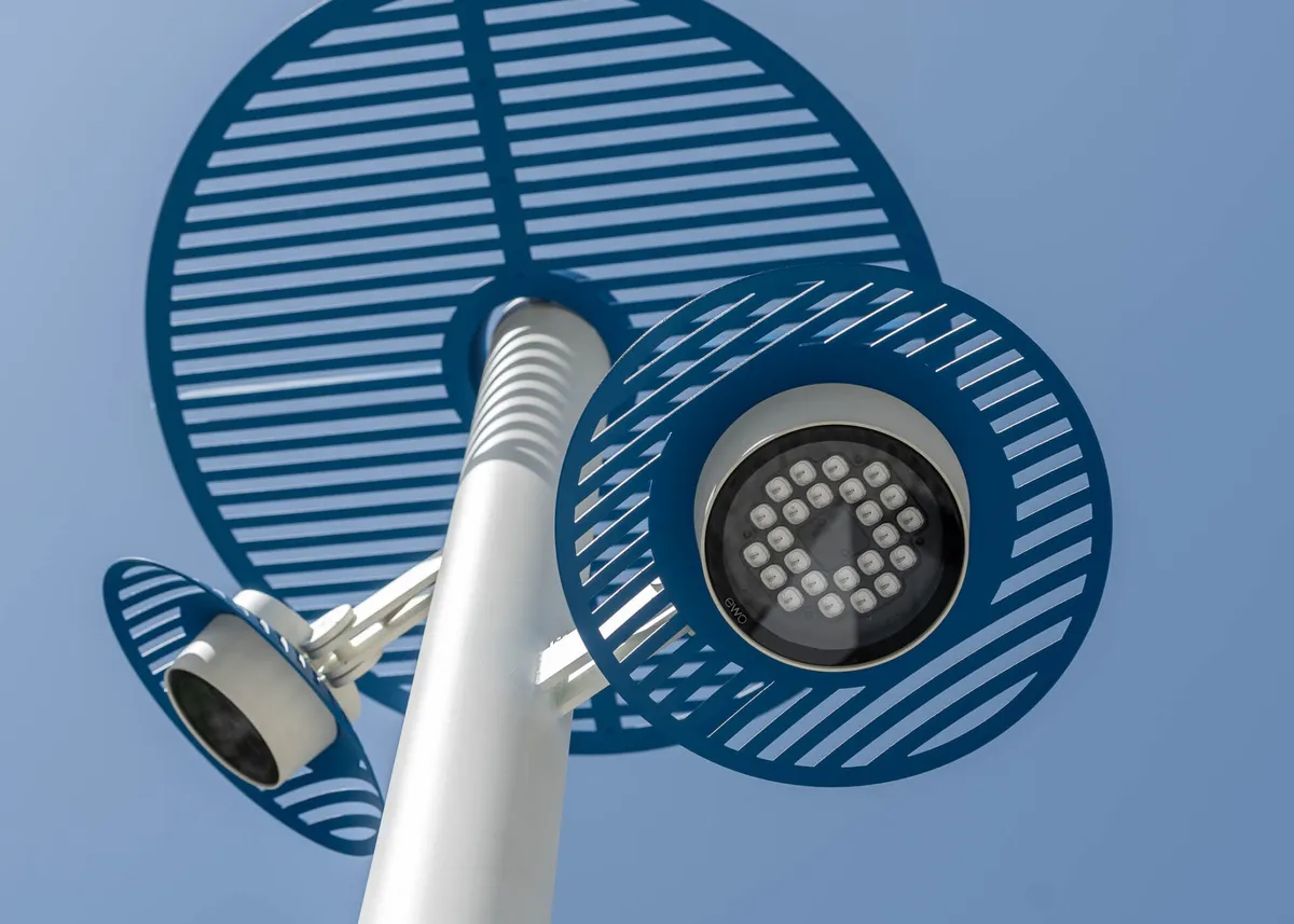 A white Chameleon pole against a blue sky, carrying two heads at different heights, with a graphic of large ringed leaf shapes drawn over the photograph.