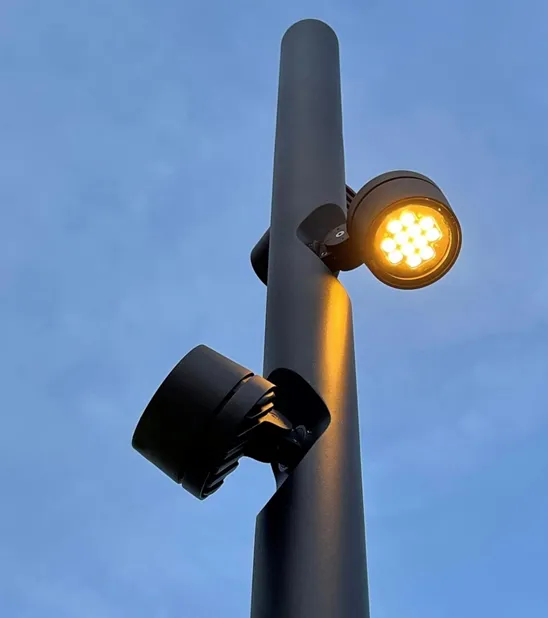 Looking up a dark Chameleon pole at dusk. Two heads point in different directions from the same column, the upper one lit warm with its cluster of LEDs visible.
