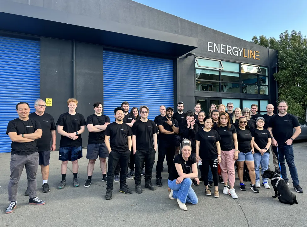 The Energyline team outside the Christchurch factory, in front of blue roller doors under the Energyline sign, most in black shirts, with a dog sitting at one end of the line.