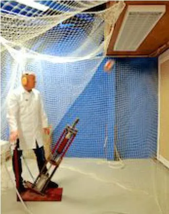 A ball impact test in progress. A launcher on a stand is aimed at a luminaire mounted high on a netted blue wall, with a dummy in a lab coat standing beside it.