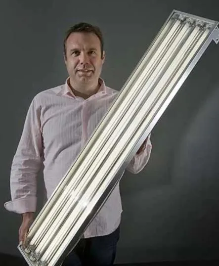 A man carrying a Revalo high bay luminaire under one arm — a long open batten holding several linear lamps in a polished reflector, about as tall as he is.