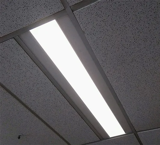 A recessed linear luminaire lit in a suspended tile ceiling, seen from below at an angle, its diffuser an even bar of white against the grey tiles.
