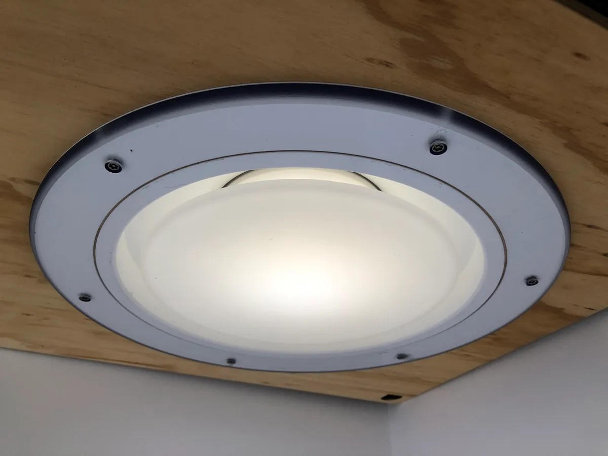 A Salvalux luminaire mounted on a plywood ceiling and lit white: a shallow dome behind a heavy bezel ring, fixed with tamper-resistant screws and no gap anywhere a ligature could be tied.