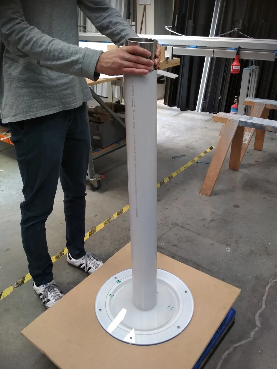 The impact test set up in the workshop: a person steadying a tall clear pipe standing upright on a base plate, with the test sample fixed underneath it.